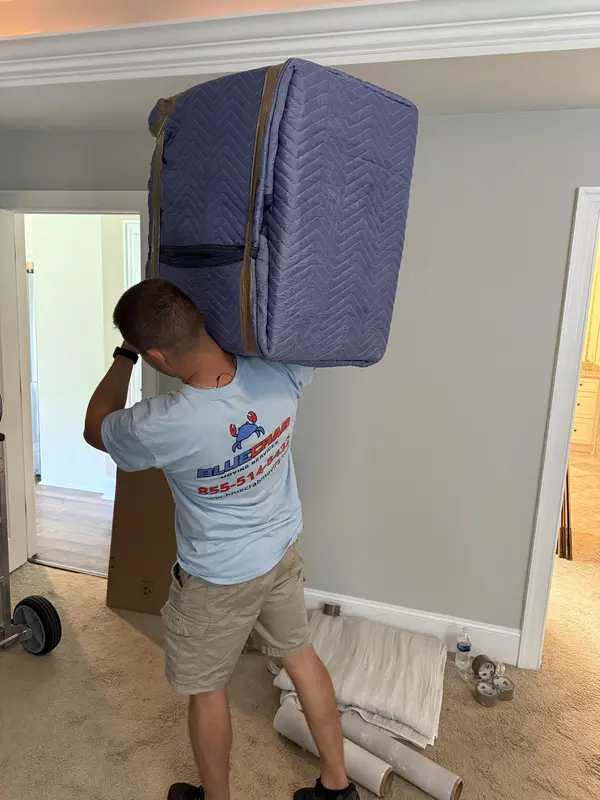 Blue Crab mover carrying a blanket-wrapped mattress out of a bedroom.